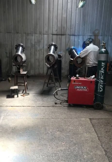 Trabajador soldando piezas de tubería en forma de Y con bridas, apoyadas sobre soportes en un taller.
